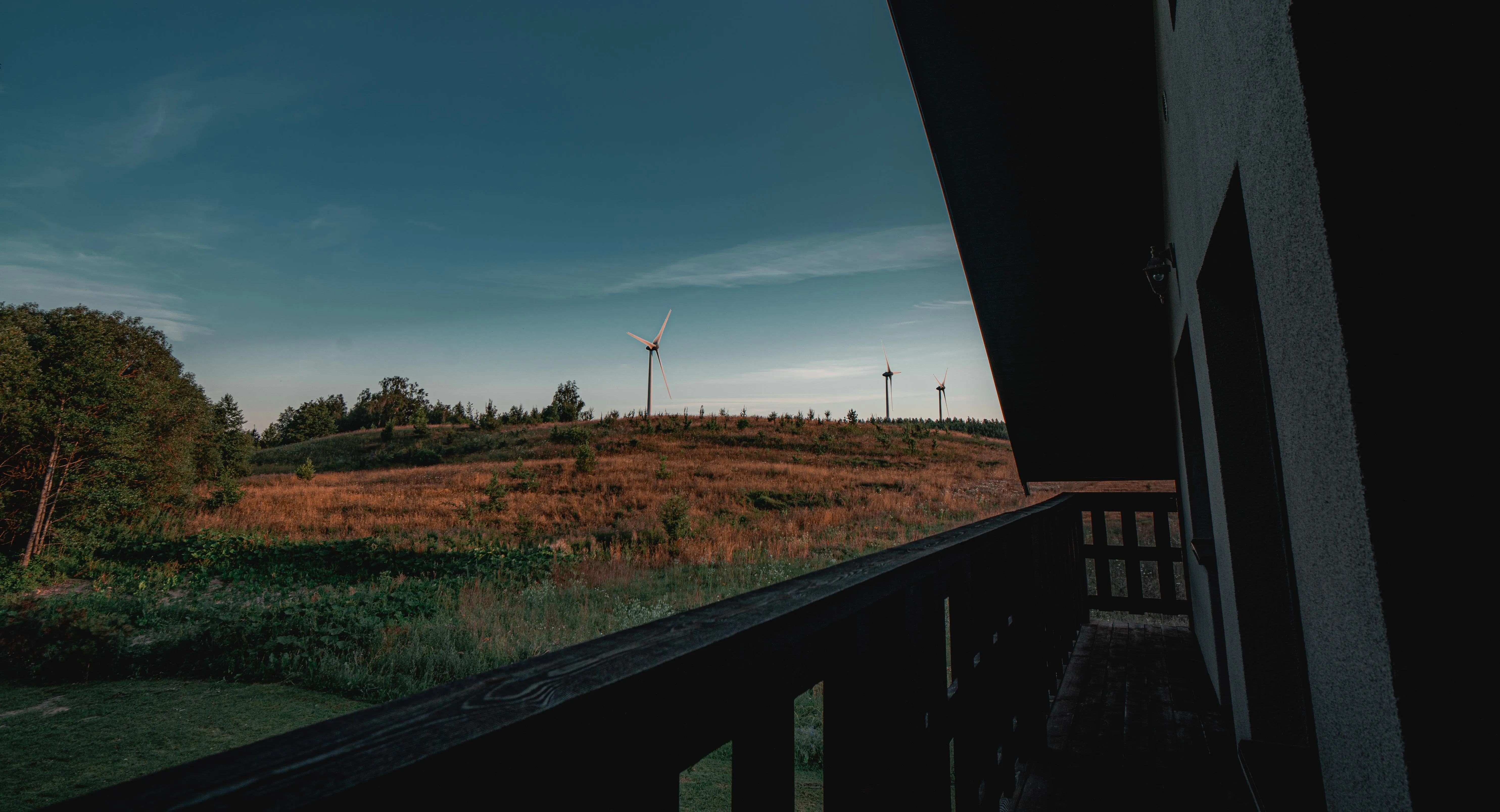 Terasa viloje — medinis balkonas, žalieji laukai, švelnus saulėlydis